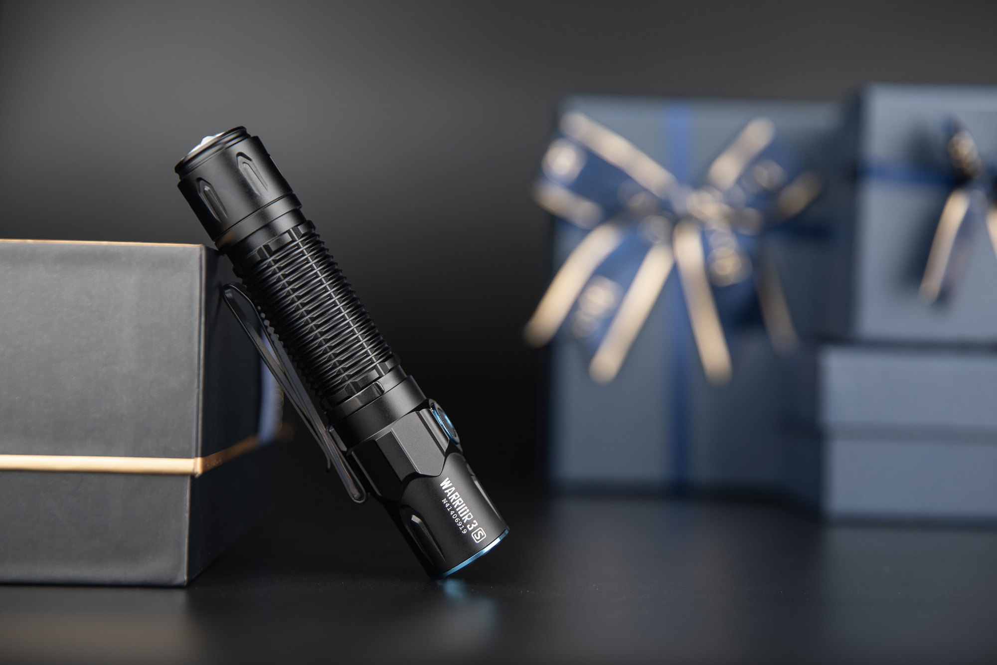 Black Olight Warrior 3S tactical flashlight leaning on a dark gift box, with blurred blue gift-wrapped boxes in the background. Perfect powerful EDC light or outdoor gear gift.
